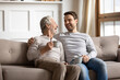 © fizkes - Overjoyed mature father and adult son drinking tea together, chatting, enjoying pleasant conversation, happy older grandfather and grandson laughing at funny joke, sitting on couch at home