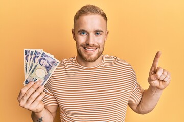 Wall Mural - Young caucasian man holding japanese yen banknotes smiling with an idea or question pointing finger with happy face, number one