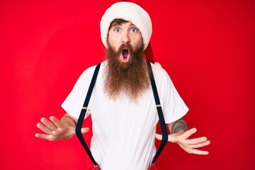 Wall Mural - Handsome young red head man with long beard wearing santa claus costume with suspenders afraid and shocked with surprise and amazed expression, fear and excited face.