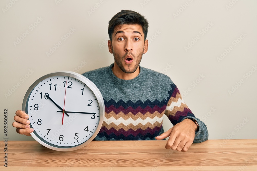 Young handsome man holding big clock scared and amazed with open mouth for surprise, disbelief face