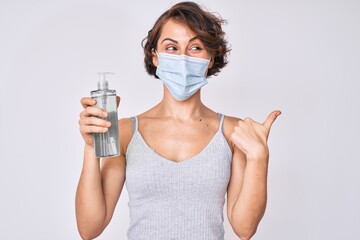 Sticker - Young hispanic woman wearing medical mask holding hand sanitizer gel pointing thumb up to the side smiling happy with open mouth