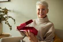 Portrait Of Stylish Charming Retired Woman With Short White Hair Sitting Comfortably On Sofa At Home Opening Birthday Present In Red Box, Looking At Camera With Happy Smile, Receiving Best Gift