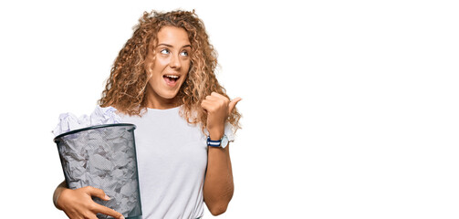 Sticker - Beautiful caucasian teenager girl holding paper bin full of crumpled papers pointing thumb up to the side smiling happy with open mouth