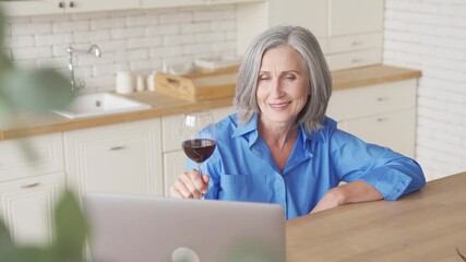 Wall Mural - Smiling older middle aged woman holding glass drinking wine video conference calling celebrating holiday party in virtual chat online meeting using laptop computer at home sitting at kitchen table.