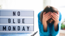 White Board With Text No Blue Monday On The Table, In The Background A Woman In Blue Clothes Depressed Near The Window,defocused.Blue Monday Day Banner Concept. Lettering.