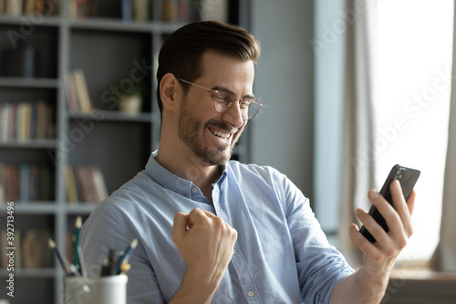 Cuadros en Lienzo Happy young man sitting at home office room holding smartphone read message feel amazed by great on-line news