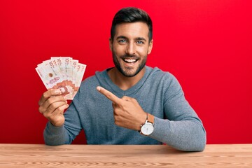 Handsome hispanic man holding colombian pesos smiling happy pointing with hand and finger