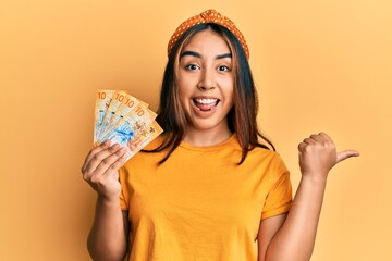 Young latin woman holding swiss franc banknotes pointing thumb up to the side smiling happy with open mouth