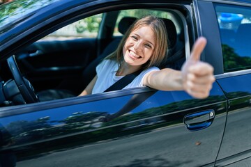 Young beautiful blonde woman smiling happy with thumb up doing ok sign sitting at the car