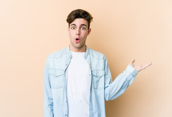 Wall Mural - Young caucasian man isolated on beige background impressed holding copy space on palm.