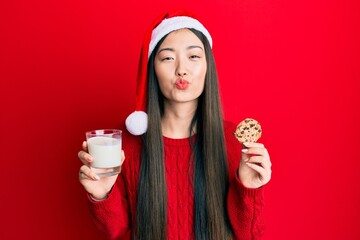 Poster - Young chinese woman wearing christmas hat holding cookies and milk looking at the camera blowing a kiss being lovely and sexy. love expression.