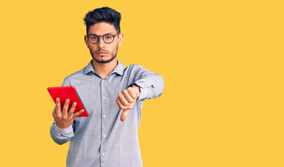 Wall Mural - Handsome latin american young man holding touchpad with angry face, negative sign showing dislike with thumbs down, rejection concept