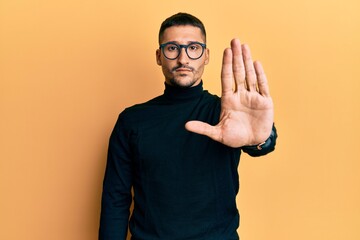 Wall Mural - Handsome man with tattoos wearing turtleneck sweater and glasses doing stop sing with palm of the hand. warning expression with negative and serious gesture on the face.