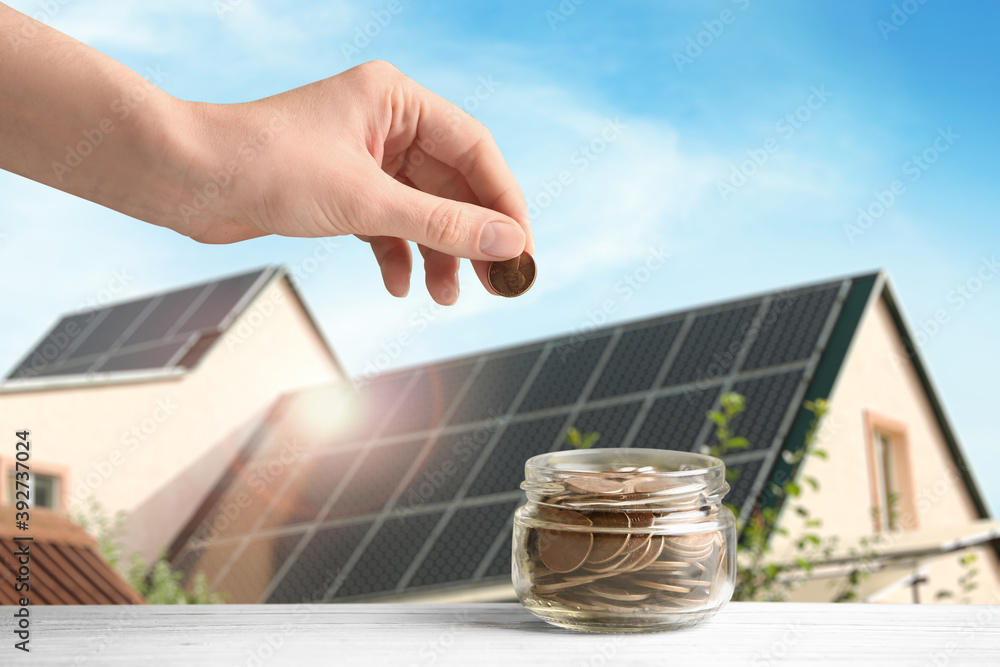 Woman putting coin into jar against house with installed solar panels on roof, closeup. Economic benefits of renewable energy - obrazy, fototapety, plakaty 