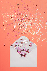 Wall Mural - Sparkling confetti pouring out of white envelope on coral background. View from above. Flat lay. Holiday, congratulation or good news concept.