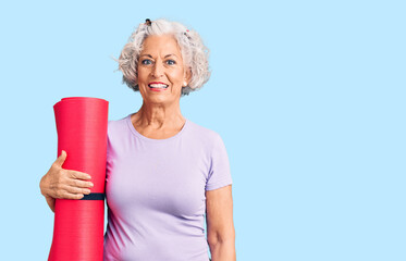 Wall Mural - Senior grey-haired woman holding yoga mat looking positive and happy standing and smiling with a confident smile showing teeth