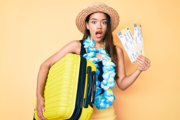 Sticker - Young beautiful latin girl wearing summer hat and hawaiian lei holding cabin bag and boarding pass afraid and shocked with surprise and amazed expression, fear and excited face.