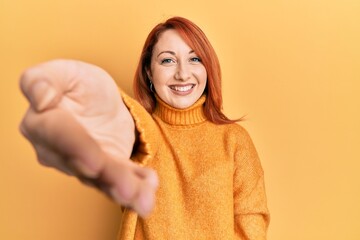 Poster - Beautiful redhead woman wearing casual winter sweater over yellow background smiling friendly offering handshake as greeting and welcoming. successful business.