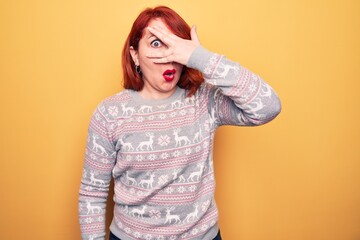 Poster - Beautiful redhead woman wearing christmas sweater with reindeer over yellow background peeking in shock covering face and eyes with hand, looking through fingers afraid