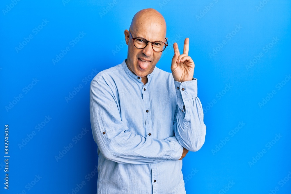 Middle age bald man wearing casual clothes and glasses smiling with happy face winking at the camera doing victory sign. number two.
