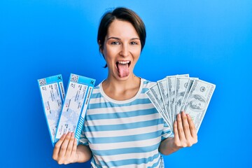 Wall Mural - Young brunette woman with short hair holding boarding pass and dollars sticking tongue out happy with funny expression.