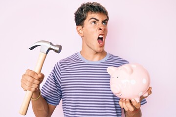 Young handsome man holding piggy bank and hammer angry and mad screaming frustrated and furious, shouting with anger. rage and aggressive concept.