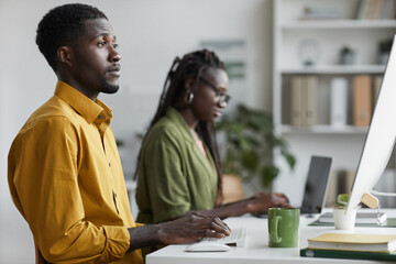 Side view portrait of young African-American man using PC in modern office and looking at computer screen, copy space