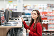 © _KUBE_ - Beautiful young woman in a red sweater, choosing a laptop. In the background, shelves with electronics. Concept of choosing and purchasing digital equipment