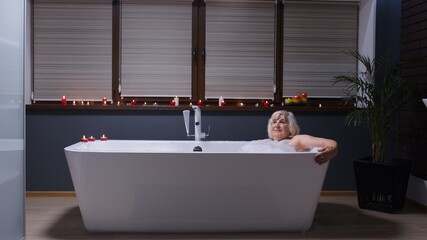 Beautiful active senior woman lying in warm bath with bubbles, enjoying relaxation. Elderly people. Side view of cheerful happy grandmother at luxury home bathroom in a romantic setting with candles