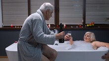 Sexy Senior Woman Grandmother Is Taking Foamy Bath In Luxury Bathroom With Candles. Elderly Grandfather Man Giving Drinking Glass With Champagne To Grandma. Honeymoon. Happy Life Of Pensioners In Love