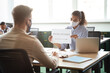 © Kostiantyn - Business as new normal. Young mixed race woman wearing protective face mask showing paper with text SOCIAL DISTANCE to male colleague while working together in office