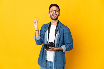 Young photographer girl isolated on yellow background smiling and showing victory sign