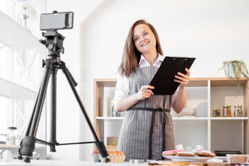 Sticker - Online conference. Webinar training. Homemade culinary. Happy chef woman with black clipboard having videochat masterclass on smartphone light home kitchen interior.