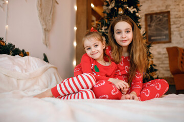 Wall Mural - Two cute little sisters in red pajamas are sitting on the bed and having fun against the background of a Christmas tree