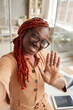 © Seventyfour - High angle portrait of young African-American woman waving at camera while taking selfie photo or live streaming from home office