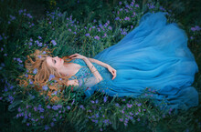 Art Photo Fairy Tale Sleeping Beauty. Fantasy Woman Lies On Blooming Meadow In Long Blue Medieval Vintage Dress. Summer Nature Background, Green Grass Bed From Purple Flowers. Girl Enchanted Princess