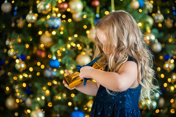 a beautiful cute little girl with blond hair in a blue dress holds a christmas present in her hands 