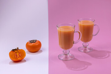 Wall Mural - two glasses of persimmon juice on a pink background and fruit on a white background