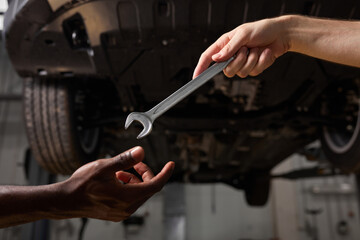 partial view of unrecognizable auto mechanics with wrench in repair shop, african and caucasian men cooperate, work as team, give instruments, tools to each other