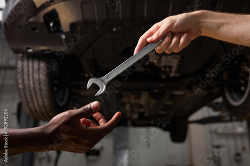 partial view of unrecognizable auto mechanics with wrench in repair shop, african and caucasian men cooperate, work as team, give instruments, tools to each other