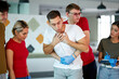 © alfa27 - young man choking with male instructor standing behind performing heimlich maneuver indoors, group of people look at them