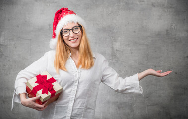 Happy beautiful blond woman wears christmas santa claus hat - cap and holding red gift box - present with ribbon, isolated on gray background