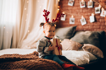 Wall Mural - A little girl holding a large gift in a paper box on a large bed. The child has antlers on his head. Christmas mood