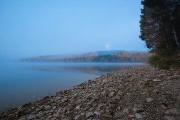 Wall Mural - Scenic view of the river banks with trees covered in autumn colored leafs and mist during sunrise and full moon set