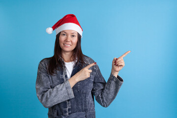 mock up smiling Asian young woman in a Santa Claus hat pointing her index fingers at an empty space for text isolated on a blue background.