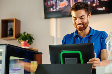 Smiling bearded coffeeshop barista working on POS terminal when entering customer order or accepting payment