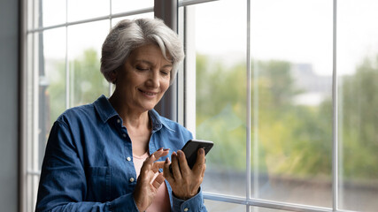 Close up smiling mature woman using smartphone, browsing apps, standing near window at home, senior grey haired female looking at phone screen, chatting or shopping online, enjoying leisure time