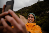 Fototapeta Panele - Attractive young male trail runner smiling wide taking photo of self on smartphone after run on mountain path in cloudy weather