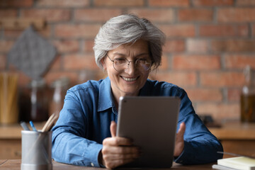 Close up smiling mature woman wearing glasses using computer tablet, chatting or shopping online, looking at screen, happy senior female having fun with device, browsing apps, making video call