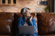 © fizkes - Thoughtful mature woman wearing glasses looking to aside, solving financial or health problems, using laptop, sitting on cozy couch alone, pensive senior female having doubts, working online